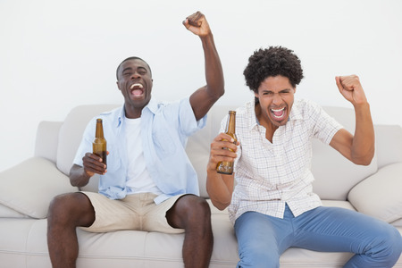 Football fans cheering holding beer bottles at home in the living roomの写真素材