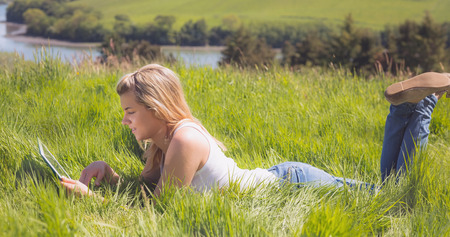 Pretty blonde lying on grass using her tablet on a sunny day in the countrysideの写真素材