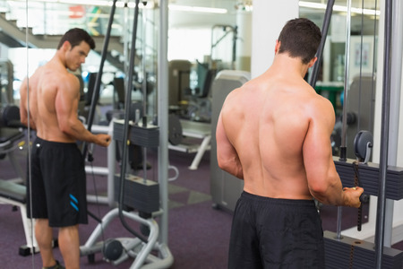 Rear view of a shirtless young muscular man using triceps pull down in gym