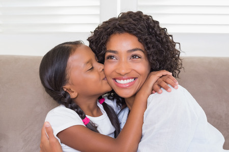 Pretty mother sitting on the couch with her daughter kissing her cheek at home in the living roomの写真素材