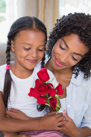 Pretty mother sitting on the couch with her daughter holding roses at home in the living roomの写真素材