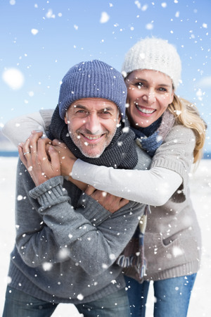 Attractive couple hugging and smiling at camera on the beach in warm clothing against snow fallingの写真素材