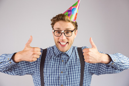 Geeky hipster wearing party hat smiling at camera on grey backgroundの写真素材