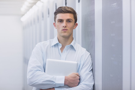 Portrait of a confident technician holding a laptop and leaning against a tower in a large data...