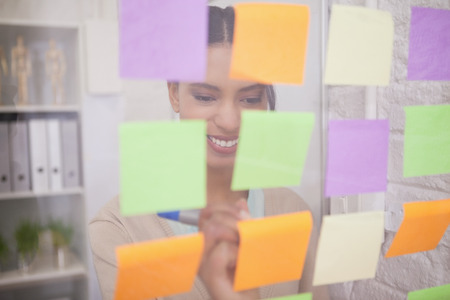 Smiling businesswoman writing on sticky notes on window in the office