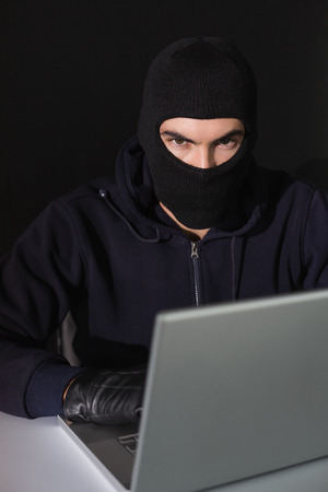 Burglar sitting hacking laptop while looking at camera on blakc backgroundの写真素材