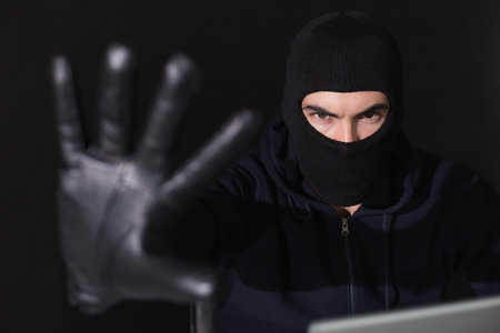 Hacker in balaclava gesturing and looking at camera on black backgroundの写真素材