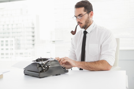 Hipster businessman using a typewriter smoking a pipe in his officeの写真素材