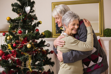Senior couple hugging beside their christmas tree at home in the living roomの写真素材