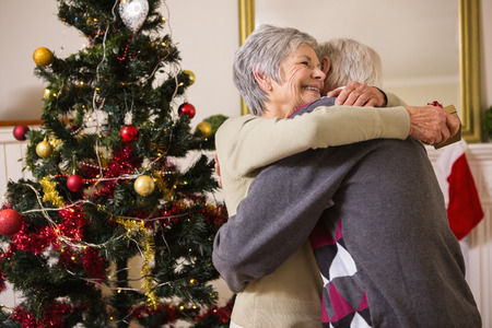 Senior couple hugging beside their christmas tree at home in the living roomの写真素材