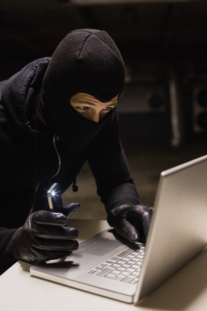 Robber hacking a laptop while making light with his phone on black backgroundの写真素材