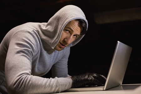 Man in hood jacket hacking a laptop while looking at camera on black backgroundの写真素材