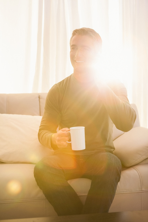 Smiling man on the phone and holding mug at home in the living roomの写真素材
