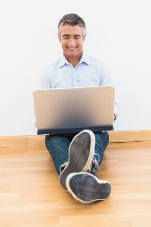 Happy man sitting on parquet using laptop at apartmentの写真素材
