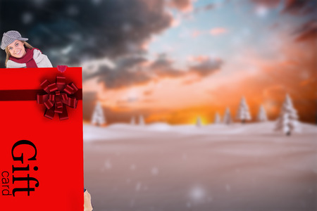 Happy blonde in winter clothes showing card against snowy landscape with fir treesの写真素材