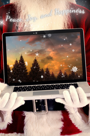 Santa Claus presents a laptop against snowy landscape with fir treesの写真素材