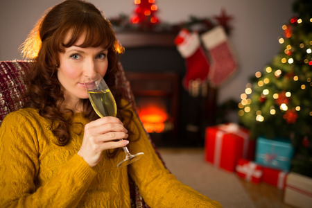 Pretty redhead drinking glass of champagne on couch at christmas at home in the living roomの写真素材