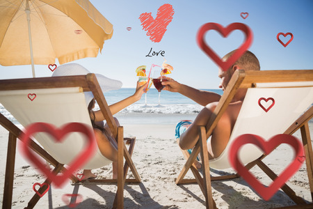 Happy couple clinking their glasses while relaxing on their deck chairs against love heartの写真素材