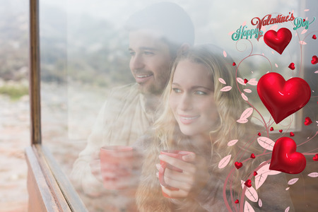Thoughtful content couple with cups looking through window against cute valentines messageの写真素材