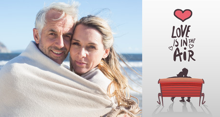 Smiling couple wrapped up in blanket on the beach against love is in the airの写真素材