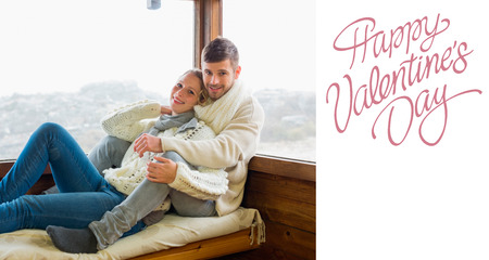 Couple in winter clothing sitting against cabin window against cute valentines messageの写真素材
