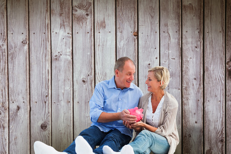 Happy mature couple holding piggy bank against digitally generated grey wooden planksの写真素材