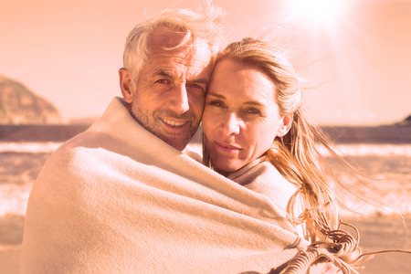 Smiling couple wrapped up in blanket on the beach on a sunny dayの写真素材