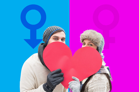 Attractive young couple in warm clothes holding red heart against pink and blueの写真素材