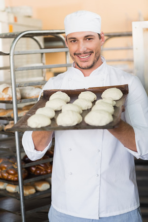 Smiling baker holding tray of raw dough in the kitchen of the bakeryの写真素材