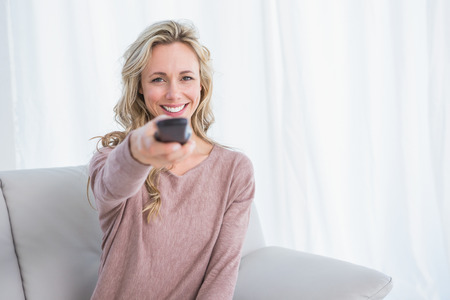 Happy blonde on couch changing tv channel at home in the living room