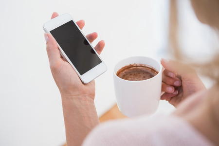 Blonde holding smartphone and mug of hot chocolate at home in the living room