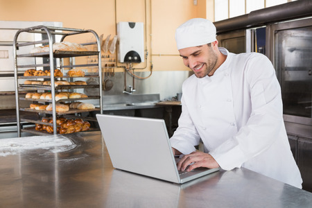 Smiling baker using laptop on worktop in the kitchen of the bakeryの写真素材