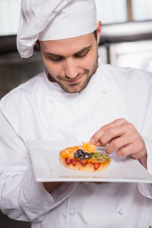 Focused baker putting flower on the pastry with fruit in the kitchen of the bakeryの写真素材