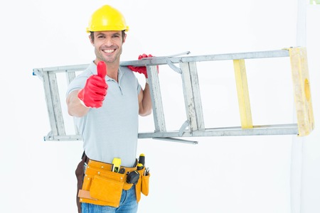Portrait of confident worker carrying step ladder while showing thumbs up over white backgroundの写真素材