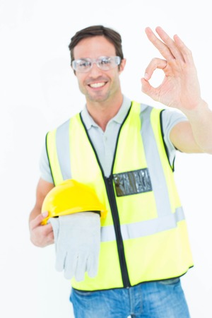 Portrait of carpenter holding gloves and hardhat while gesturing OK sign against while background