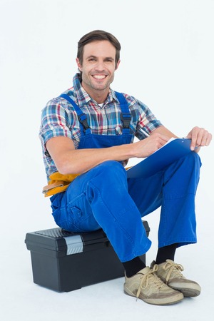 Portrait of happy plumber writing notes while sitting on tool box over white backgroundの写真素材