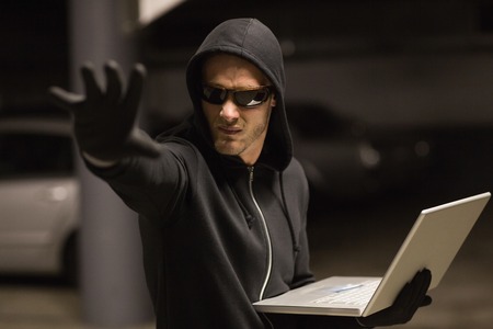 Hacker in balaclava gesturing and using laptop on shadowy background