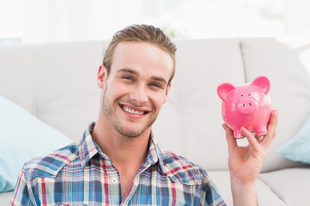 Smiling man showing pink piggy bank at home in the living roomの写真素材