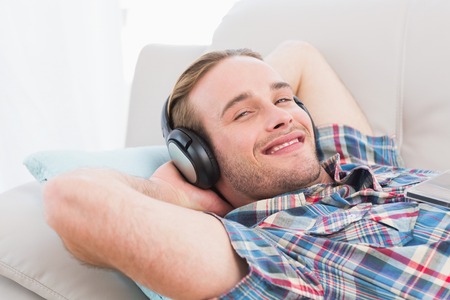 Smiling man lying on sofa listening music at home in the living roomの写真素材