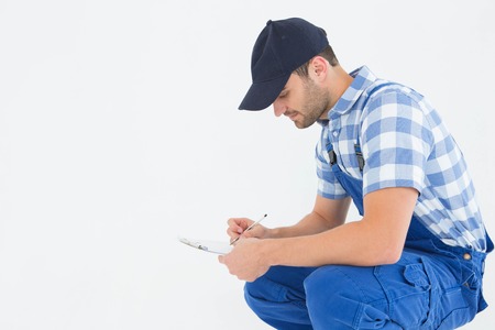 Side view of young handyman crouching while writing on clipboard on white background