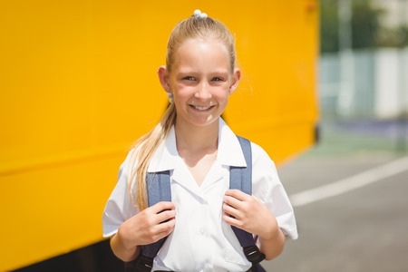 Cute pupil smiling at camera by the school bus outside the elementary school