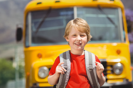 Cute pupil smiling at camera by the school bus outside the elementary school