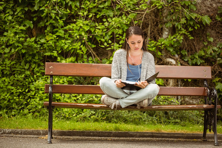 Student sitting on bench listening music with mobile phone and revising in park at school
