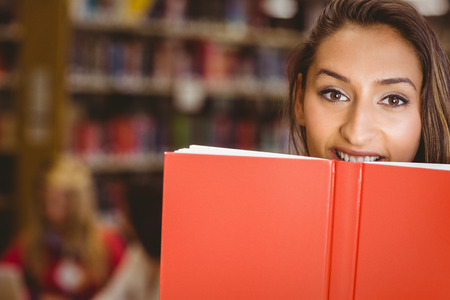 Pretty brunette student holding book in front of her face in library
