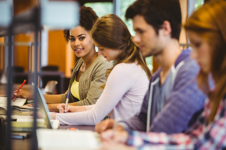 Student looking at camera while studying with classmates in libraryの写真素材