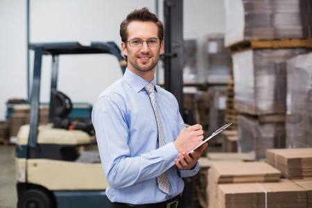 Manager looking at camera while holding clipboard in a large warehouseの写真素材