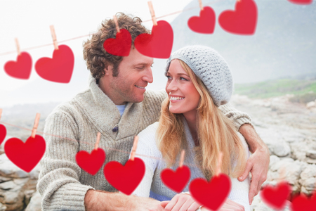 Happy couple sitting together on rocky landscape against hearts hanging on a lineの写真素材