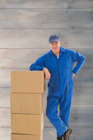 Happy delivery man leaning on pile of cardboard boxes against pale grey wooden planksの写真素材