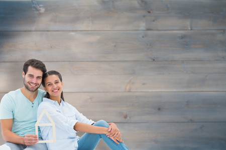 Cute couple sitting holding a house shape against bleached wooden planks backgroundの写真素材