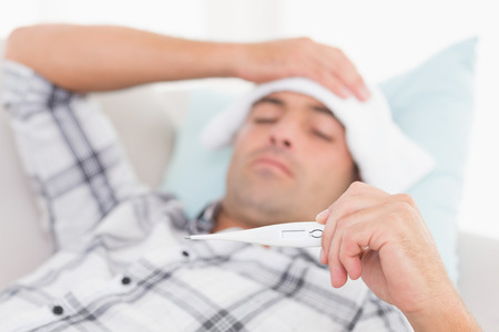 Sick man checking temperature on thermometer while lying on sofa at home